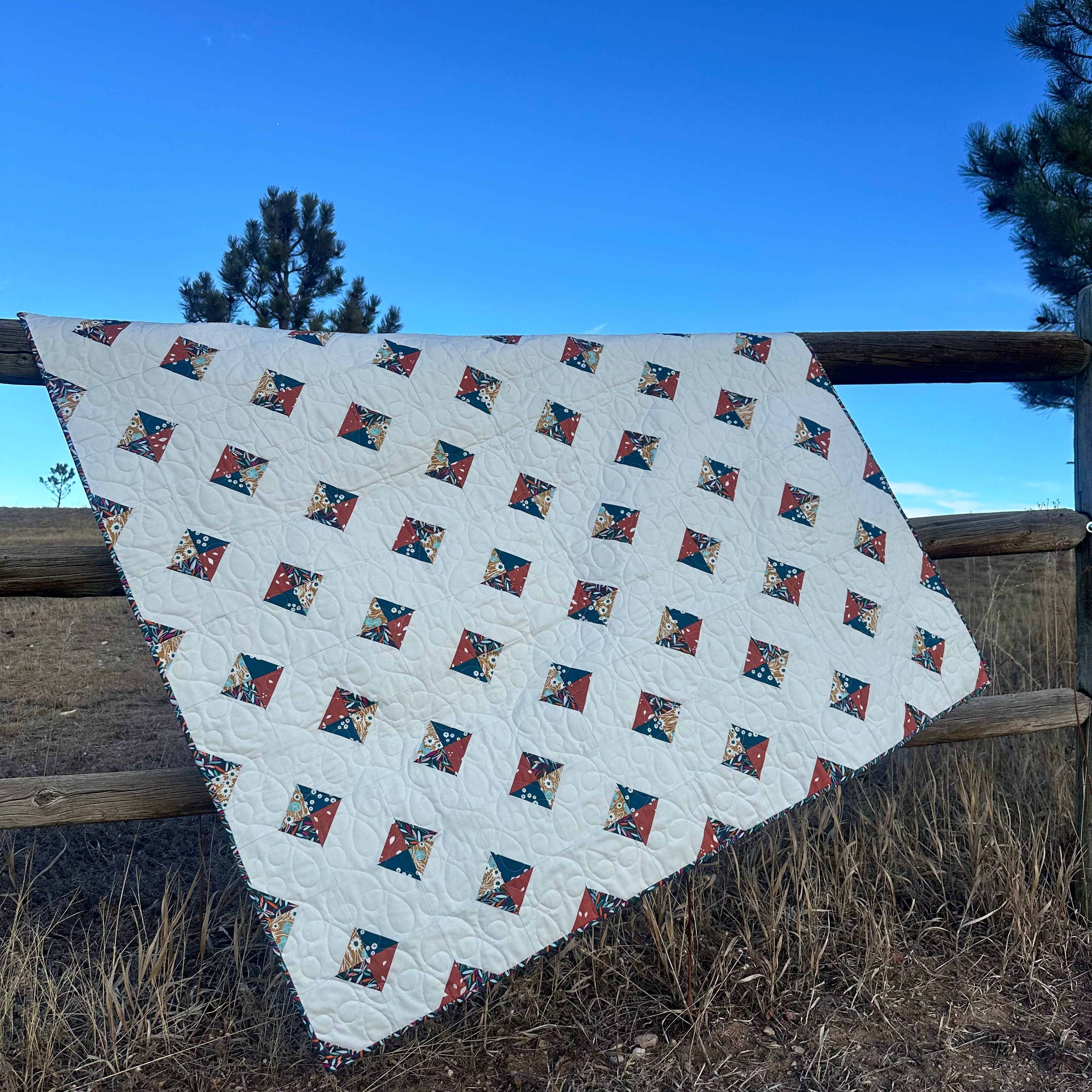 Quilt with geometric pattern draped over a wooden fence against a blue sky